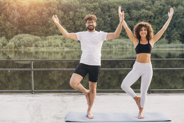 Obraz premium Happy couple doing yoga tree pose on a mat by a lake, enjoying nature and balance during a fitness workout