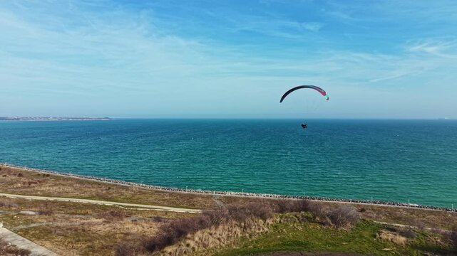 A paraglider is flying high above the ocean on a sunny day. The coast is visible in the background with calm water and clear skies. People may be enjoying the beach below.