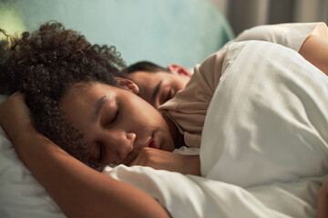Young adult Black woman and young adult Caucasian man sleeping closely together in bed, both...