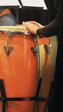 Close up of a percussionist's hands on a vibrant orange conga drum. Warm stage lighting highlights the instrument and the artist during an energetic live music performance at a jazz festival