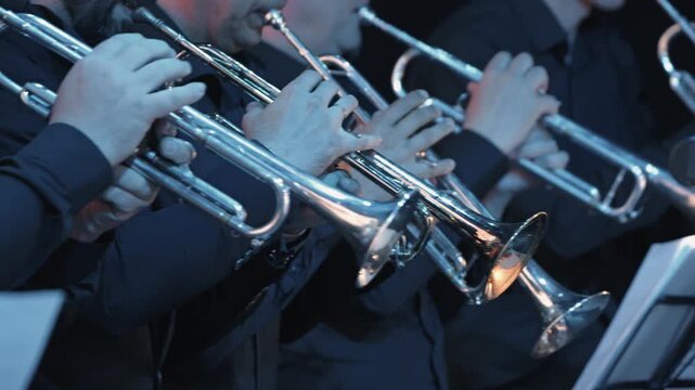 A close-up view of the trumpet section of a jazz band performing live on stage. Musicians in dark formal wear hold their silver instruments, with warm stage lights reflecting on the polished metal
