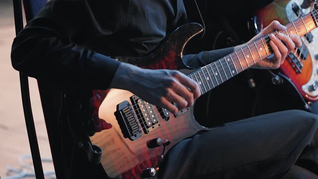 Close-up of a male guitarist's hands playing chords on a six-string electric guitar. The musician is performing on stage at a concert, with soft lighting highlighting the instrument and his skill