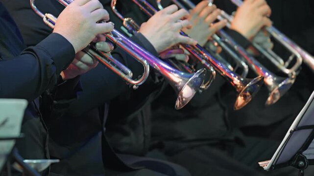 Close-up of a trumpet section performing on stage. Musicians in dark attire hold shiny instruments reflecting colorful stage lights