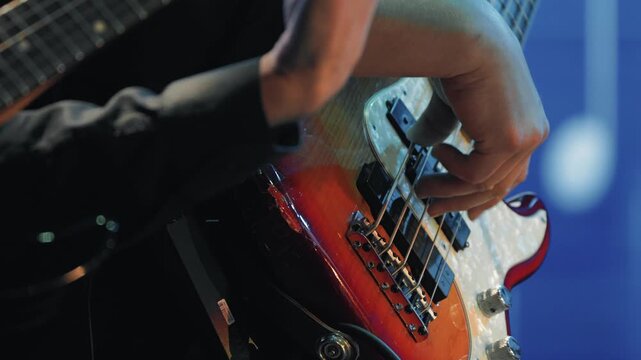 A tight shot of a musician's hands playing a sunburst bass guitar. The scene is lit with warm stage lights against a blue background, capturing the energy of a live jazz performance