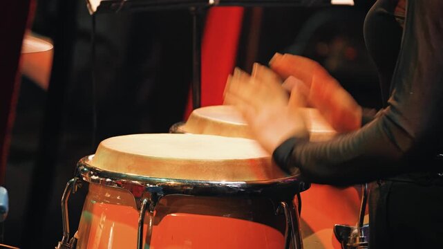 Dynamic close-up of a musician's hands playing conga drums on stage. The warm, atmospheric lighting highlights the orange drums creates an energetic mood for a live jazz latin music event