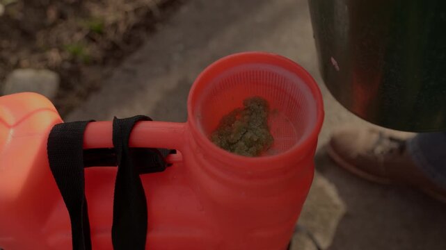 Close-up of a gardener pouring water into an orange backpack sprayer, dissolving granular fertilizer to create a liquid solution for treating plants in the garden during a sunny day