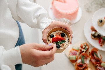 Female hands holding a paper cup with creamy dessert, fresh strawberries, and blueberries. A pink frosted cake and party appetizers are in the background. © AndreyZayats