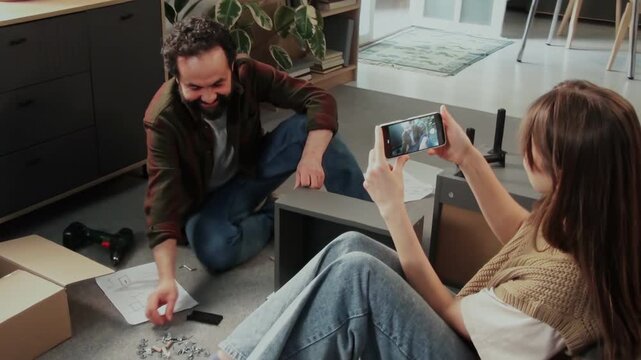 Handheld zoom camcorder shot of Caucasian woman sitting on floor at home, filming husband assembling flatpack cabinet on smartphone, chatting while enjoying happy moments of family life
