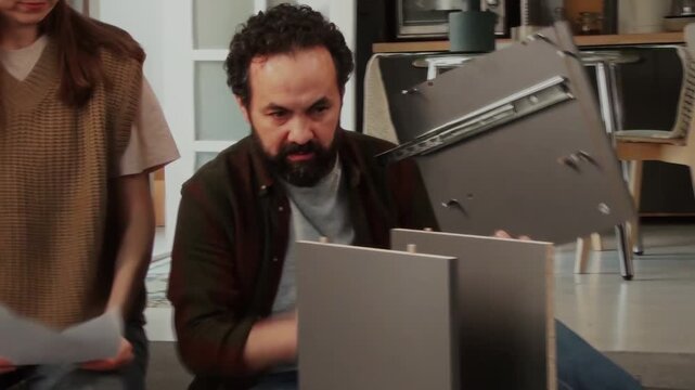 Zoom handheld camcorder shot of busy mature Caucasian couple assembling cabinet together at home, woman holding instruction manual and giving advice while man is attaching panel with fasteners