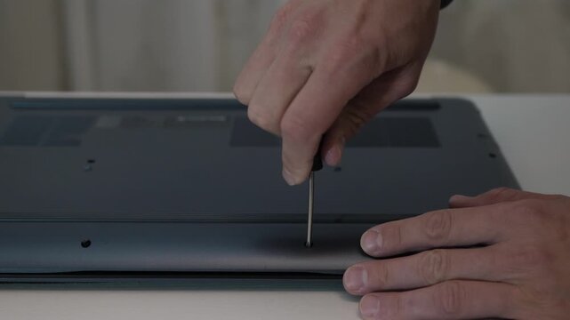 Closeup hands unscrewing laptop screws, technician loosening bottom cover with screwdriver on neutral table, slow careful motions, repair process