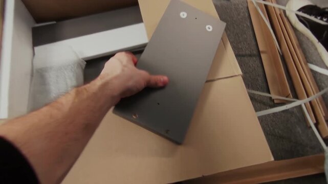 Handheld UGC shot of man filming assembly of flatpack drawer with partner at home, male hand holding instruction manual, taking fiberboard panels out of box, capturing daily family life on camcorder