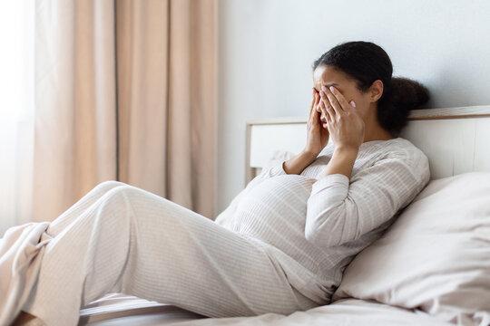 Pregnant Black woman lying in bed covering her face with hands, expressing stress or headache. Emotional fatigue during pregnancy concept in bedroom