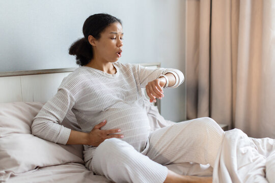 Pregnant Black woman sitting on bed timing contractions, checking watch with focused expression. Labor preparation concept in bright cozy bedroom