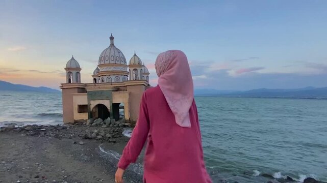 Muslim woman in a hijab stands by the shore, looking at the iconic damaged Floating Mosque of Palu, Central Sulawesi, a symbol of resilience after the 2018 tsunami.