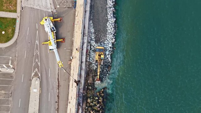 Workers are managing construction tasks at a waterfront site where a crane lifts heavy materials while an excavator digs near the water's edge in clear weather.