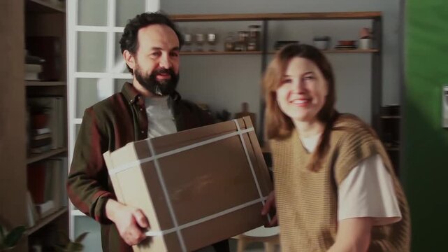 Medium handheld shot of mature Caucasian husband and wife standing in living room at home, holding large heavy box with flatpack drawer, discussing purchase, filmed on camcorder for family memory