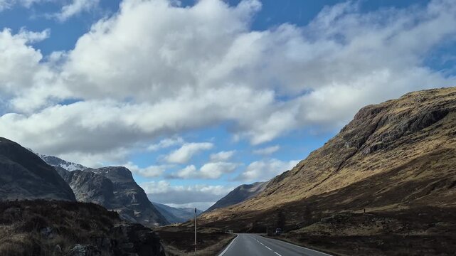 Driving asphalt road leading into the mountain pass of Glencoe, peaks in the Scottish Highlands, sunny day with clouds, nature travel destination road trip, 4K