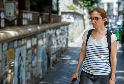 Woman tourist wearing a striped shirt and glasses, carrying a backpack, walking along the iconic muretto di alassio adorned with ceramic tiles, exploring the charm of alassio