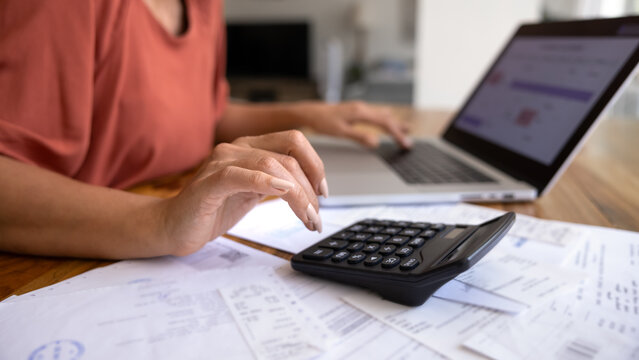 Close-up of female managing personal finances at home, using calculator and modern laptop surrounded by bills and receipts, as symbol of budgeting, accounting, and family expenses control. Accountancy
