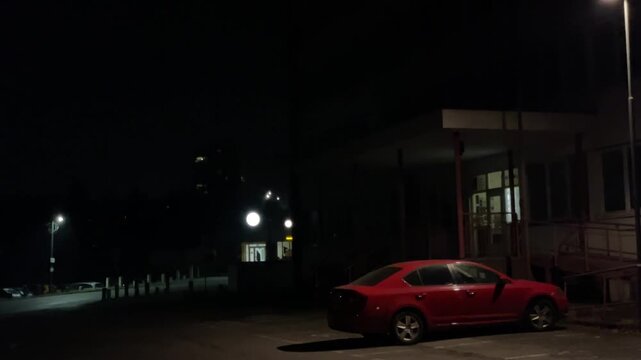 Night parking lot with red car, dim lamppost casting long shadows across asphalt, closed storefront and quiet street, distant headlights