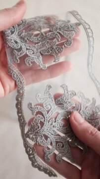 Close-up of hands holding and inspecting a delicate silver-grey embroidered lace trim with a floral damask pattern on a fine mesh background. Perfect for fashion design, and high-end texttile