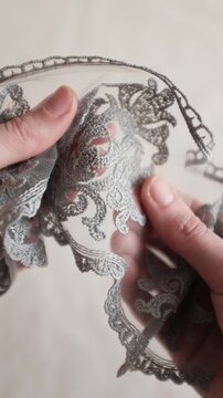 Close-up of hands holding and inspecting a delicate silver-grey embroidered lace trim with a floral damask pattern on a fine mesh background. Perfect for fashion design, and high-end texttile