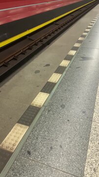 Vertical video of underground or subway. Blue LED safety line along metro platform floor, granite and concrete tiles, tactile markers and approaching train lights create guarded