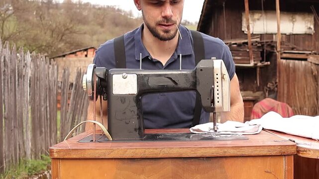 Country style man with hat, suspenders and straw in mouth sitting on a chair, pulling it closer and preparing to start sewing on a vintage machine in a rustic outdoor setting.