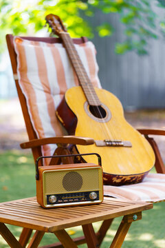 Cozy outdoor relaxation space with wooden garden chair, acoustic guitar and vintage radio placed on small table. Peaceful backyard leisure scene for music, rest and summer recreation in green garden.