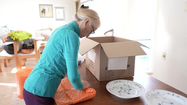 Mature woman carefully wraps and packs family heirloom china in a box for shipping to her family member while at home.