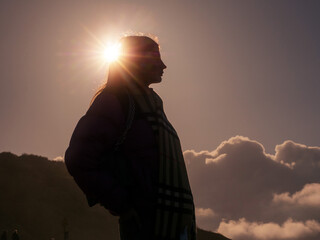 Silhouette stands against radiant sun glow. Soft clouds drift behind distant hills. Person gazes upward, quiet and thoughtful. Golden light frames their profile in warmth © mark_gusev