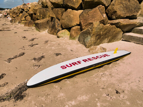 Sunlit surf rescue surfboard rests on sandy beach. Yellow fin and red lettering stand out against white deck. No people present, making it ideal for calm, quiet moments. Life guard tool.