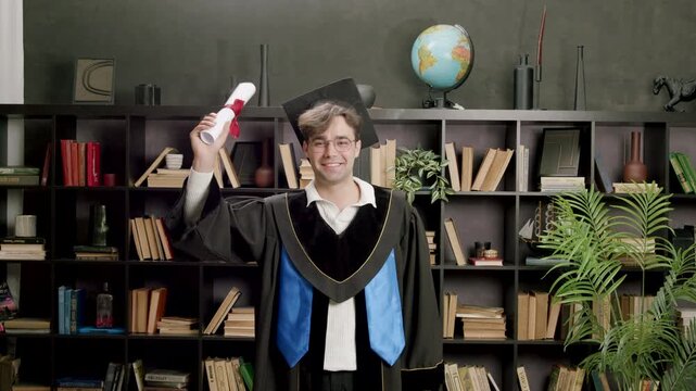 Male graduate in black cap and gown proudly holds diploma in front of bookshelf filled with books and decorative items in a modern indoor setting