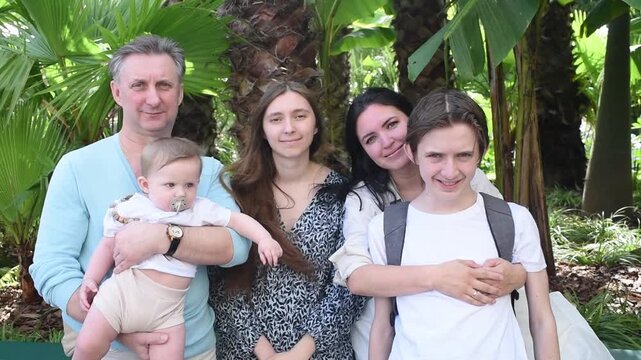 Happy multigenerational family of five posing under palm trees. Middle-aged father holding infant son, mother with teenage son and adult daughter smiling and looking at camera.