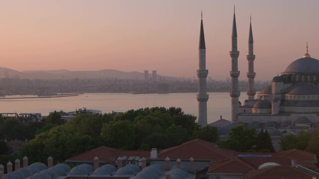 A wide view of Istanbul skyline and waterfront with tall mosque minarets rising above green trees. Soft sunset light reflects on the water while city buildings stretch across the horizon.