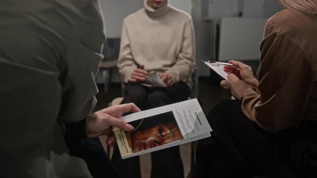 Detail shot of unrecognizable woman participating in support group meeting for women and listening to therapist speaking while sitting on chair in circle, focus on brochure in hand