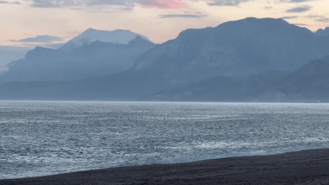 Coastal view of the Mediterranean Sea with the Taurus Mountains in the background. Hazy sunset sky, dark pebble beach in the foreground. Misty mountain peaks.