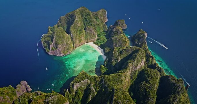 Phi Phi Island Maya Bay aerial: towering limestone cliffs draped in lush green, pristine white sand and vibrant turquoise water with boats cruising coast. Summer vacation paradise, travel to Thailand