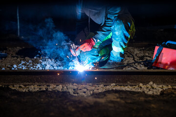 Fototapeta premium A worker in reflective gear arc welds a steel rail at night on ballast, sparks and blue smoke rise, portable welder nearby, task lighting isolates gloves and stripes.