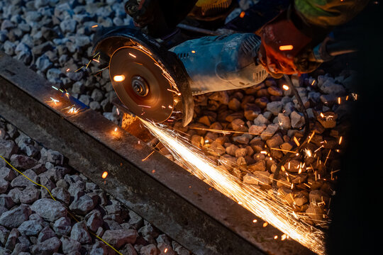 A worker in gloves operates an angle grinder on a steel railway track at night, sending orange sparks across crushed stone ballast, showing precision and safety.