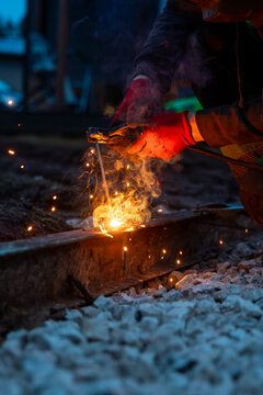 A worker in red gloves performs thermite welding on a steel rail at night, molten metal flares and sparks scatter on ballast, smoke swirls, shallow depth of field shows heat.