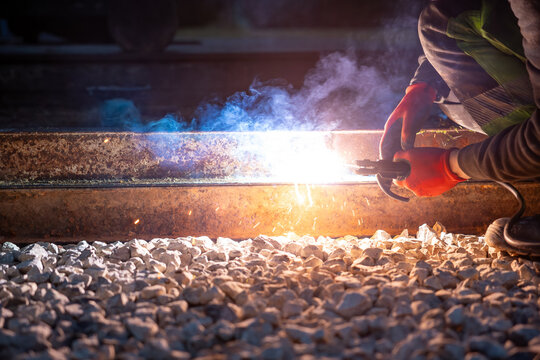 A worker in red gloves performs thermite welding on steel railroad tracks, sparks fly and blue white smoke rises over crushed ballast, ground level view at night.