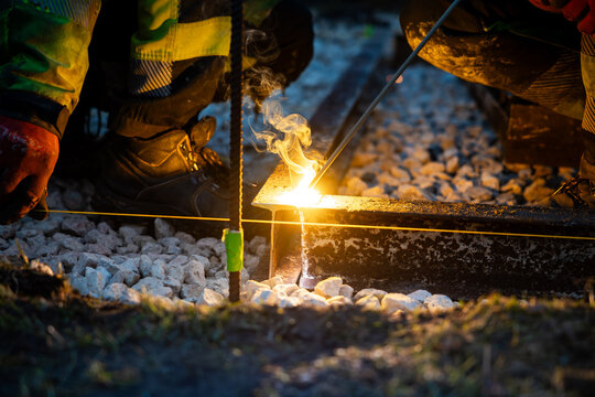 Two railway workers in high visibility gear perform a thermite weld on a steel rail, sparks and white smoke rising, guide line taut, after dusk on ballast stones.