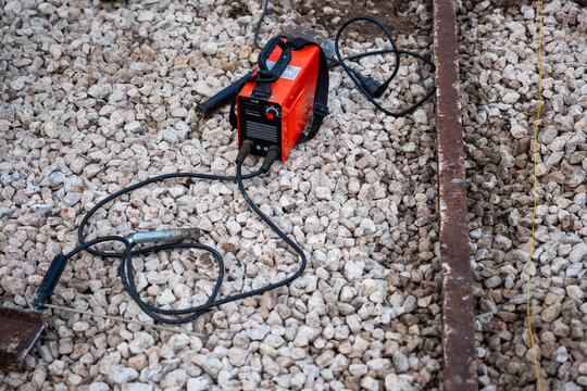 Compact orange welder rests on coarse gravel beside a rusted steel beam and a taut guide line. Daylight reveals scuffed metal and dusty cables, suggesting active field work.