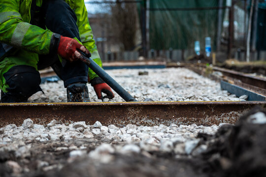 A worker in a green jacket and red gloves kneels by exposed rails, prying steel with a crowbar as crushed stone ballast extends. Tool cases and fencing sit in overcast light.