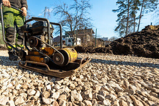 A worker in green pants runs a yellow plate compactor on crushed stone, in a fenced suburban lot with detached houses, leafless trees, soil piles, and crisp daylight.