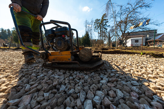 A laborer in high visibility pants operates a yellow plate compactor on coarse gravel at a rural homestead with a caravan and laundry line under midday light.