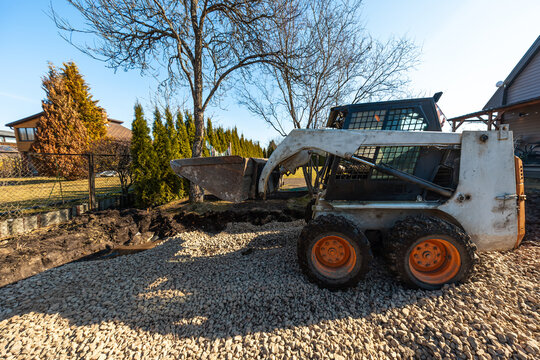 A compact skid steer with orange wheels raises a bucket on a gravel bed near a residential fence, arborvitae, and leafless trees, grading and excavating in cool daylight.