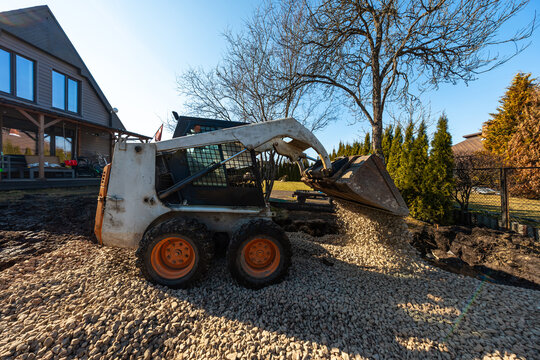 A compact skid steer pours light crushed stone onto a yard beside a modern wood sided house, with chain link fence, tall evergreens, bare branches, and crisp afternoon shadows.