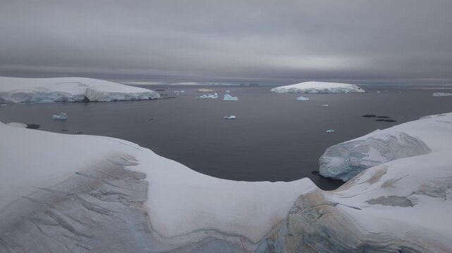 Cinematic aerial moving past the towering, blue-layered ice walls of a massive shelf in Antarctica, revealing ancient glacial strata and deep crevasses under a moody polar sky